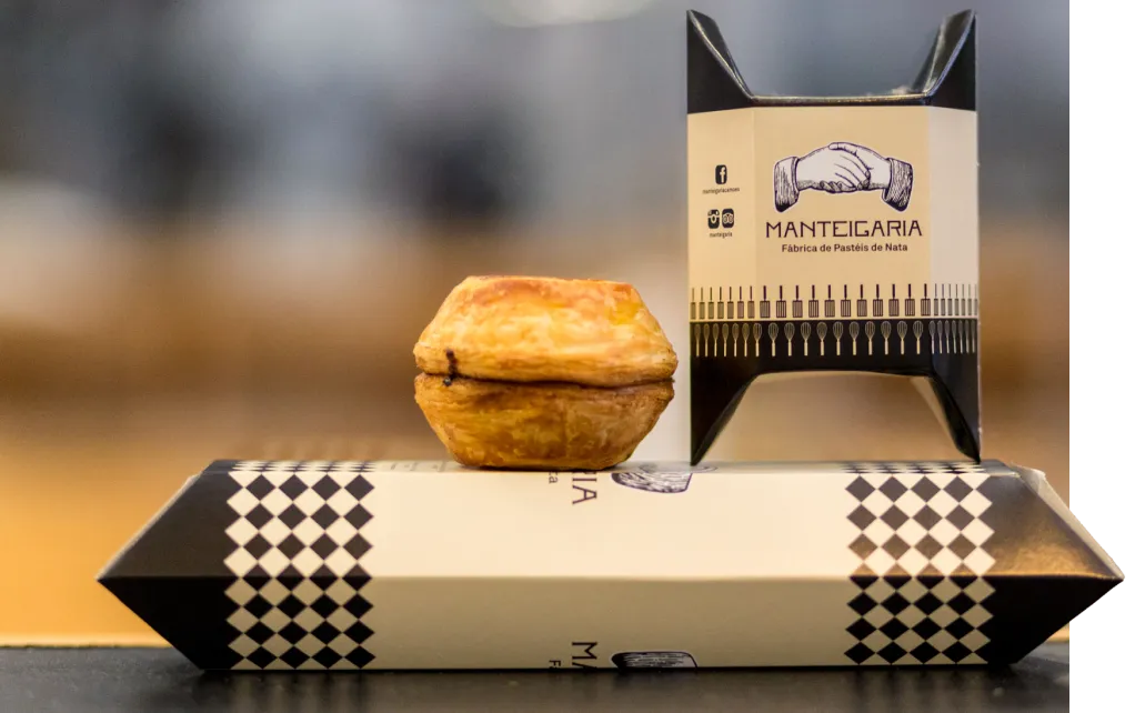 Pastel de nata sobre uma embalagem Manteigaria, com o logótipo da marca ao fundo