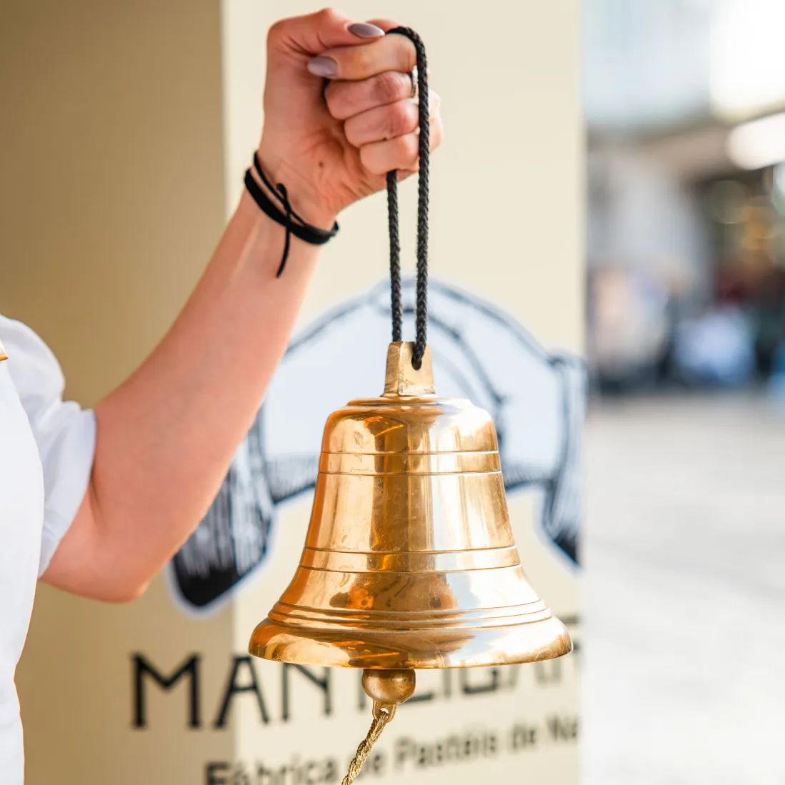 Mão a tocar o sino dourado da Manteigaria junto à fachada da loja