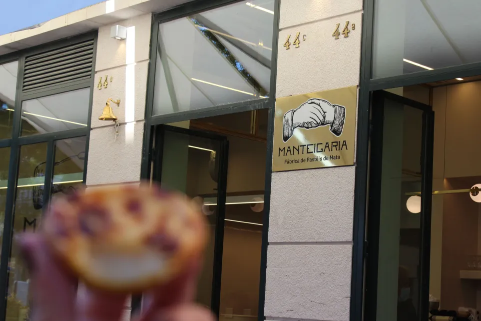 Mão a segurar um pastel de nata junto à placa da Manteigaria em Alvalade
