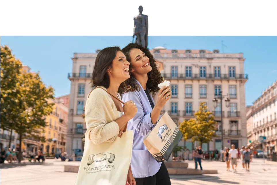 Duas mulheres a rir junto a um monumento numa praça de Lisboa, com sacos e uma embalagem da Manteigaria