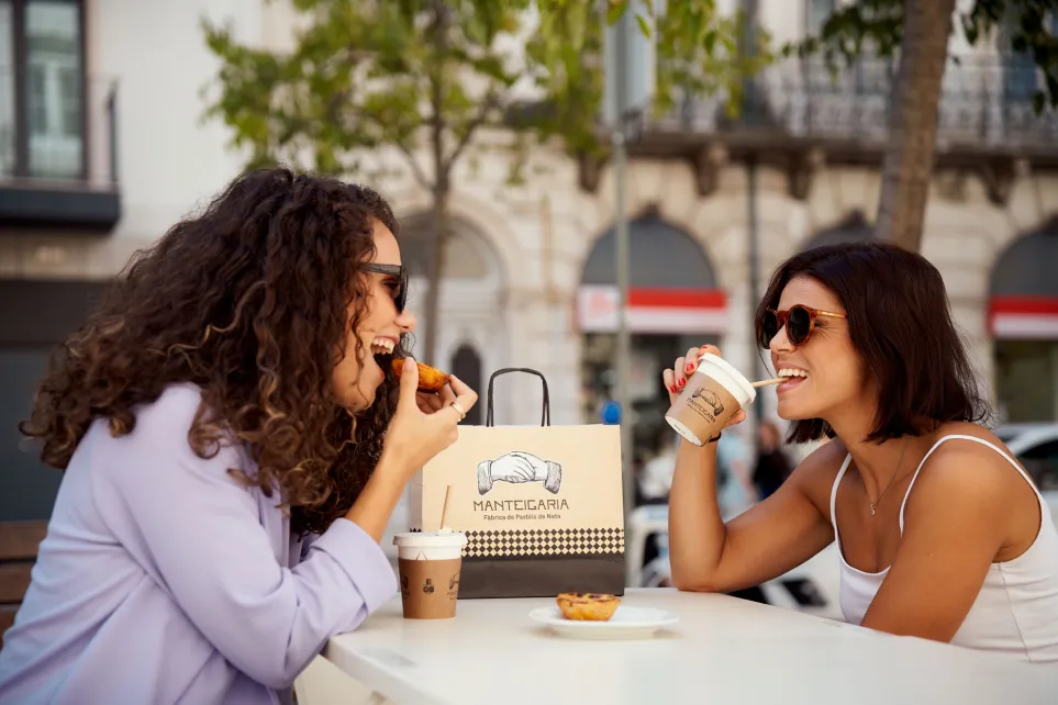 Duas mulheres a rir enquanto comem pastéis de nata numa esplanada, com um saco Manteigaria na mesa