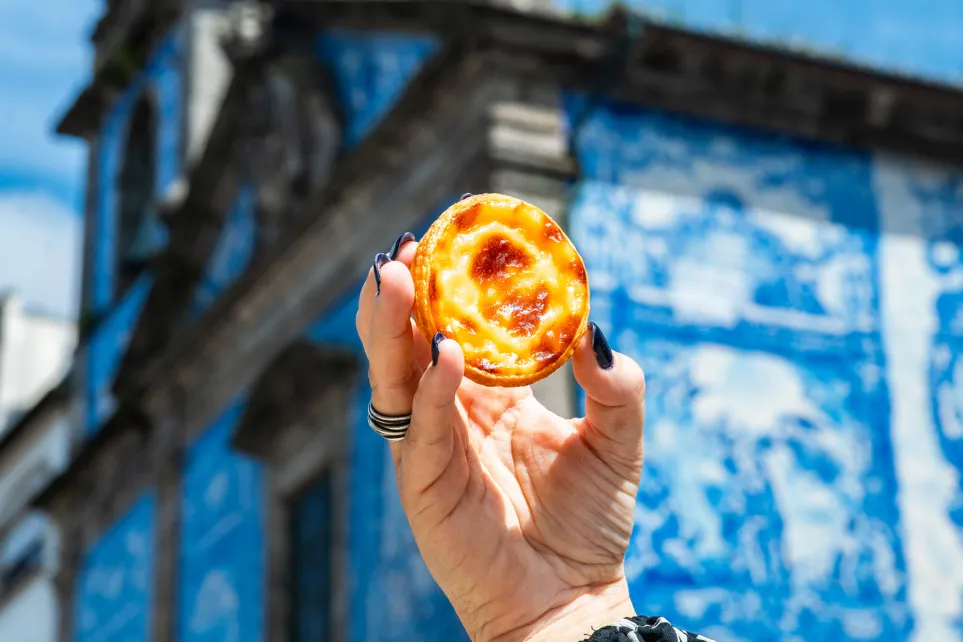 Hand holding a pastel de nata in front of a blue-tiled façade