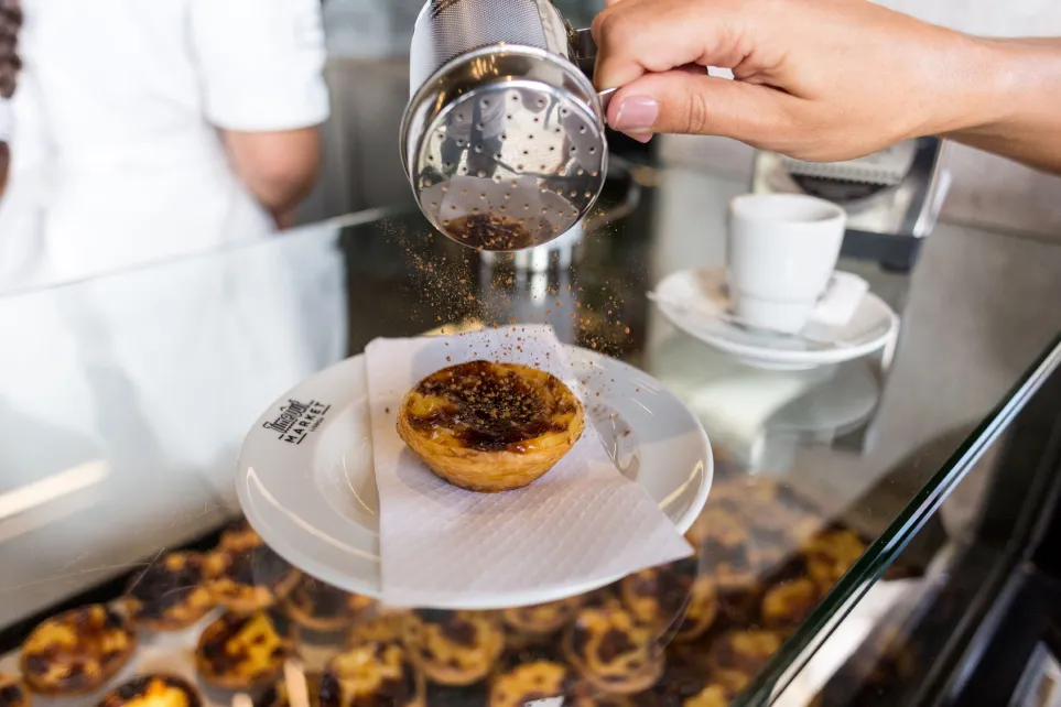Main saupoudrant de la cannelle sur un pastel de nata, dans une assiette du Time Out Market