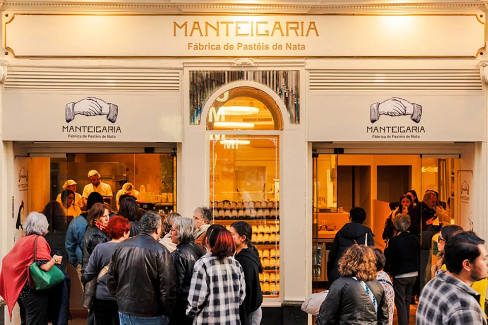 Fachada iluminada da Manteigaria à noite, com clientes em fila à entrada