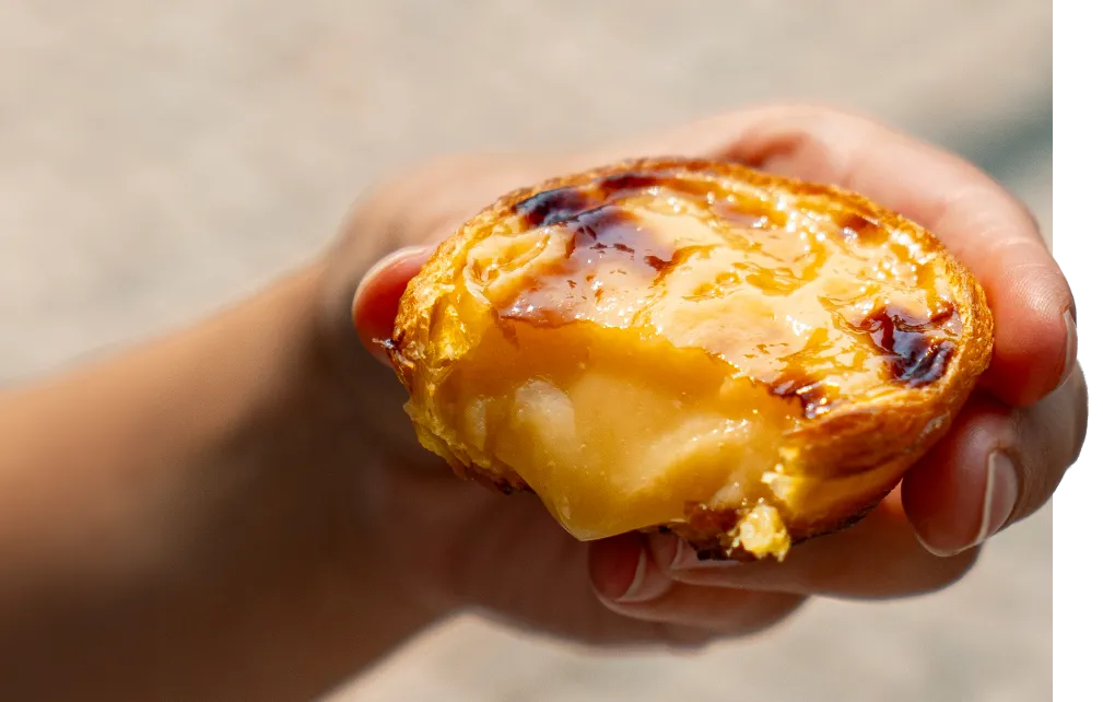 Mão a segurar um pastel de nata mordido, com o recheio cremoso e a massa estaladiça à vista