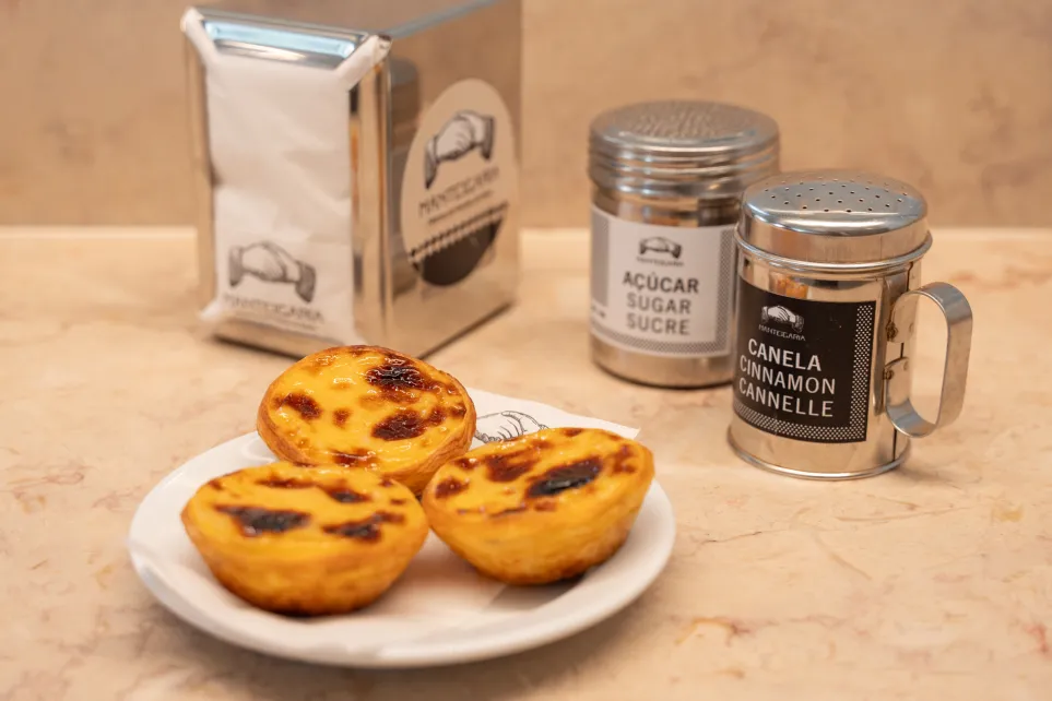 Pastéis de nata num prato Manteigaria, com latas de açúcar e canela ao lado