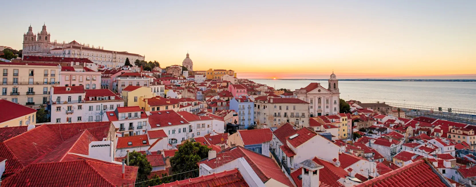 Vista do Miradouro das Portas do Sol em Lisboa ao nascer do sol, com telhados vermelhos e o rio Tejo