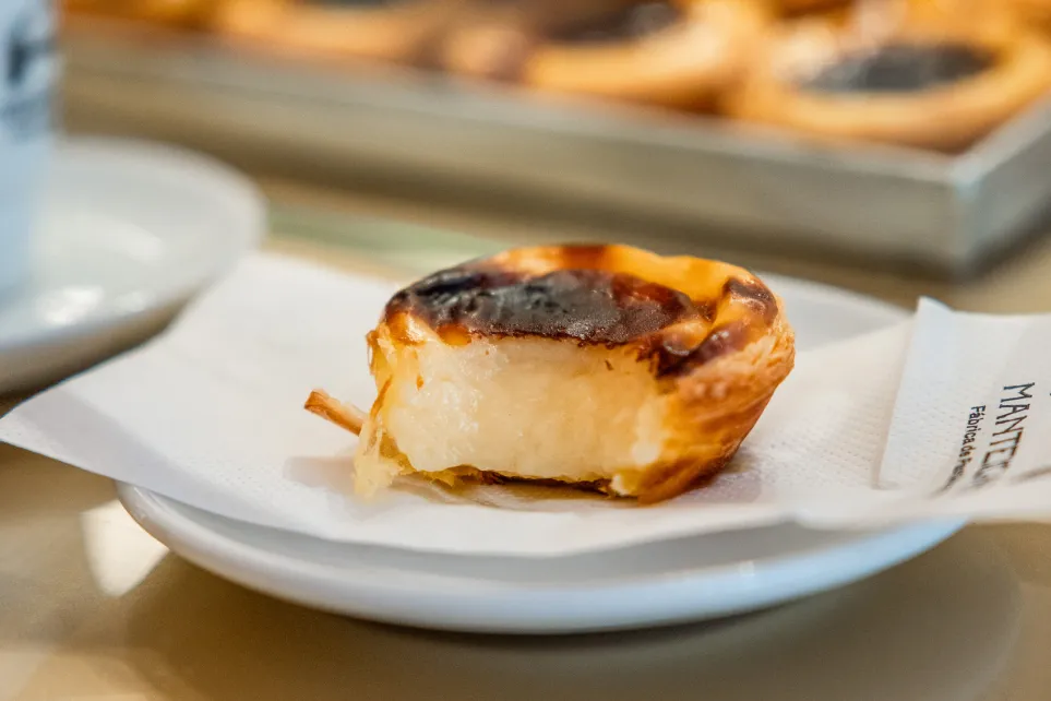 Pastel de nata num guardanapo Manteigaria, com tabuleiro de pastéis desfocado ao fundo