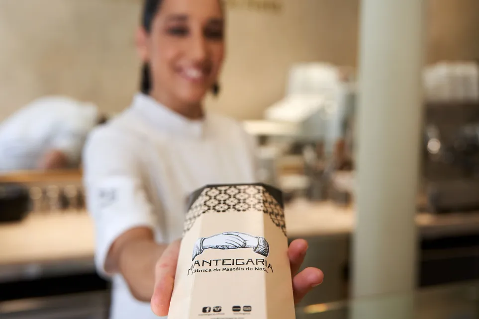 Funcionária da Manteigaria, fardada de branco, a entregar uma embalagem de pastéis de nata sorrindo para a câmara