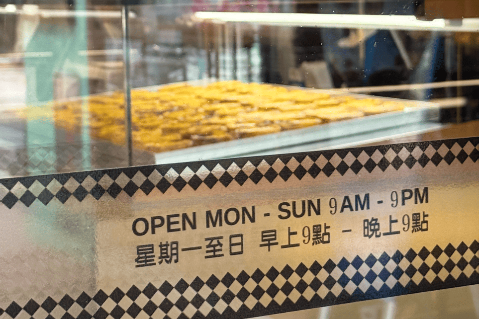 Tabuleiro de pastéis de nata na montra, com horário de funcionamento em inglês e chinês