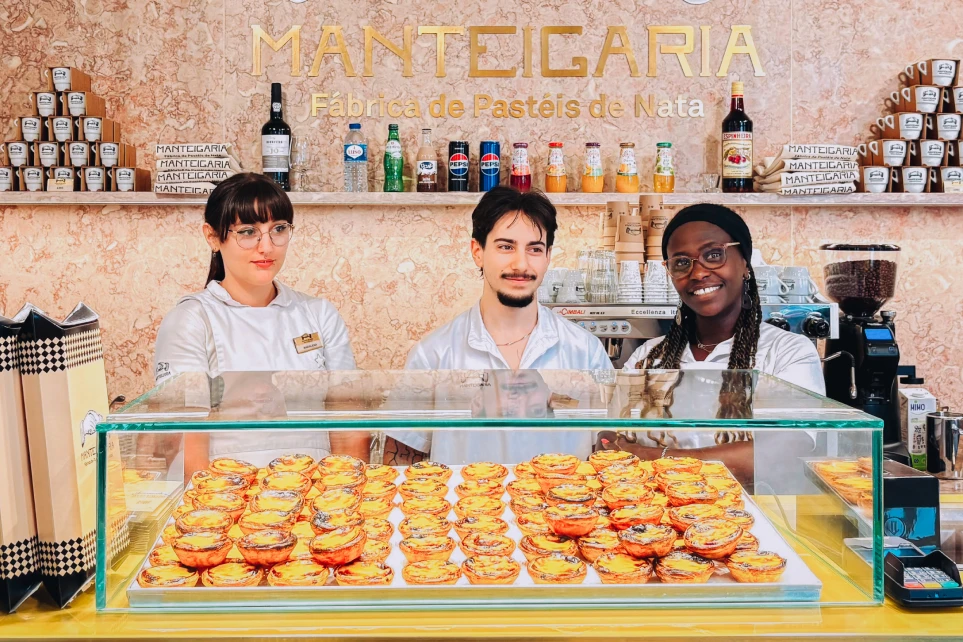 Tres empleadas de la Manteigaria sonriendo tras el mostrador, con pasteles de nata expuestos