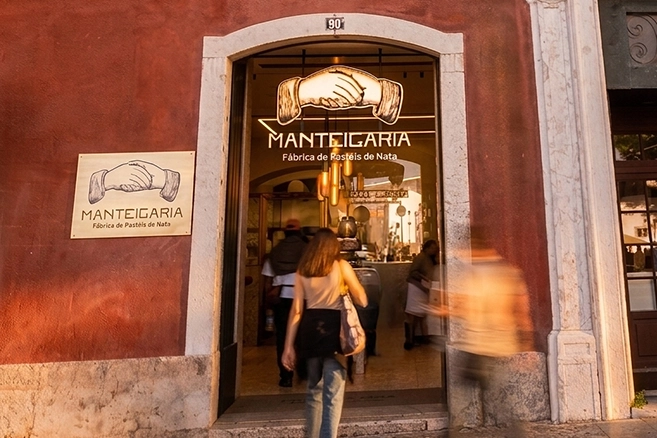 Entrance to Manteigaria on a red façade at dusk, with a person walking by